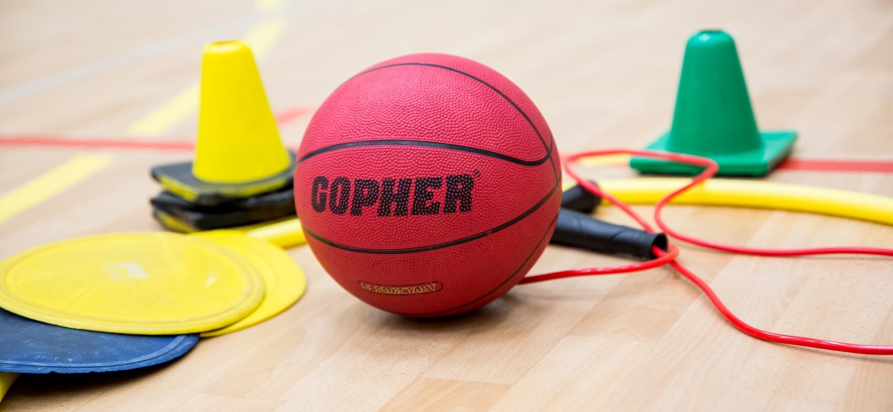 Equipment for a physical education class, including cones, a basketball and sliders, sit on the floor of a gymnasium
